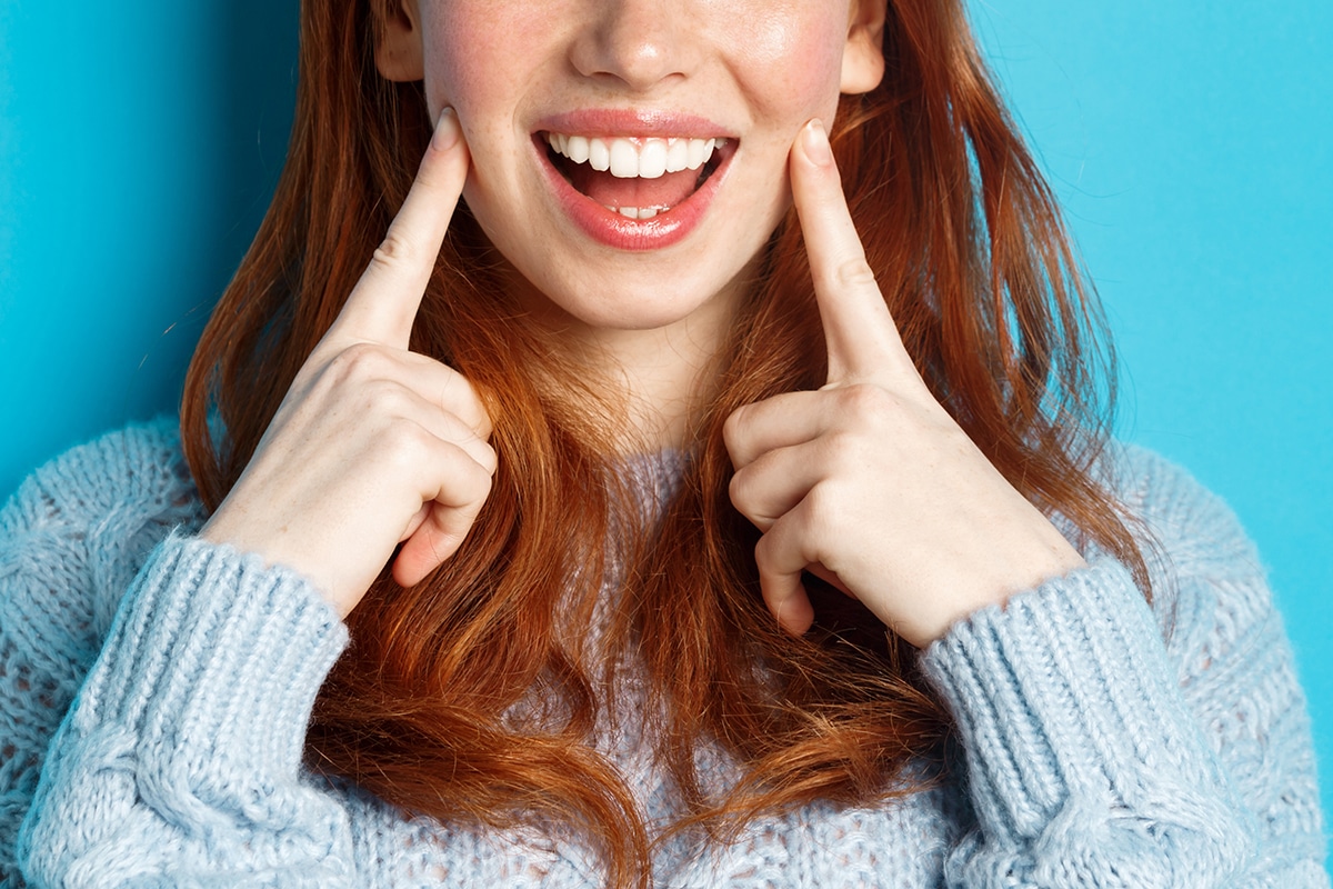 Close-up of cheerful teenage girl with red hair and freckles, poking cheeks, showing dimples and smiling with white teeth, standing over blue background
